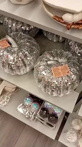 The video shows a display of disco ball pumpkins on shelves in a store. The pumpkins are covered in small mirrored squares, reflecting light and creating a sparkly effect. Some pumpkins are larger and sit on the top shelf, while smaller decorative orbs are visible on the lower shelves. The lighting is bright, highlighting the reflective surfaces of the pumpkins. The video pans across the display, showing multiple pumpkins from different angles.