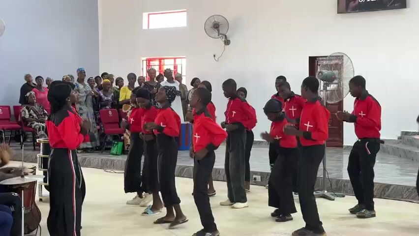 A group of young people, primarily male, are performing in a brightly lit room. They are dressed in red and black uniforms and are dancing and singing in front of an audience of seated adults. Some of the performers are wearing crosses on their shirts. To the left, a drummer plays an instrument, while another person watches. The room has light-colored walls and several ceiling fans are visible.