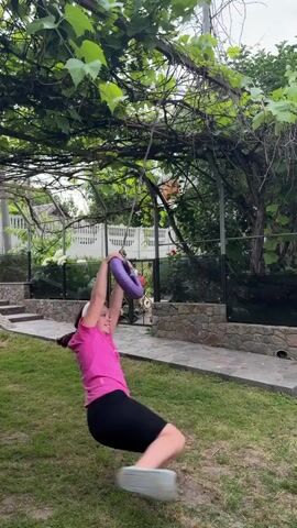 A young person in a bright pink t-shirt and black shorts is swinging on a circular purple swing suspended from a vine-covered pergola. The swing appears to be made from a hula hoop. The person is holding onto the swing with both hands and is mid-swing, with their legs bent. The background shows a grassy yard with a stone pathway and a fence. The lighting suggests it is daytime.