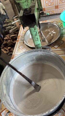 Here's a description of the video:

The video shows a kitchen setup where a machine is mixing a brown liquid in a large metal bowl. The machine is green and appears old, with a metal arm extending into the bowl and spinning. Next to the machine is a much larger pot filled with a light brown liquid, a wooden paddle is inserted into the light brown liquid. Firewood is stacked on the table nearby. People are present in the background, moving around in what seems to be a food preparation area. The table is covered with a patterned tile surface.
