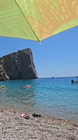 The video shows a sunny day at a pebble beach with clear turquoise water. A large, rocky cliff juts out into the sea on the left side of the frame. Several people are in the water, some on inflatable floats and others swimming. A boat is visible in the distance. On the beach, a few pairs of flip-flops are scattered on the pebbles. An umbrella, partially visible at the top of the frame, provides shade. The camera appears to be positioned under the umbrella, looking out towards the sea.