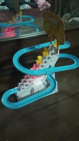 The video shows a toy set featuring a miniature roller coaster for small plastic ducks. The toy is primarily white, with a blue track winding around a staircase-like structure. The stairs are illuminated with alternating red and white lights. At the top of the stairs is a cardboard sign that reads "Small Duck" with an image of yellow ducks. Several small yellow plastic ducks are moving along the track, going up the stairs and then down the spiraling track. The background is somewhat blurred, suggesting a room with clothing and a mirror visible.