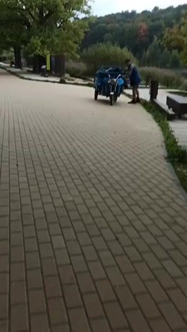 The video shows a person standing next to a blue three-wheeled vehicle with a cargo trailer attached. The vehicle is parked on a brick pathway. In the background, there is a wooden boardwalk, a sandy area, a body of water, and trees on a hillside. The person is wearing a blue jacket and shorts and appears to be interacting with the vehicle. The environment suggests an outdoor recreational area, possibly a park or a beach. The sky is overcast.