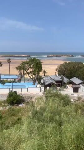 The video presents a scenic, panoramic view of a coastal landscape in Loualidia, Morocco, under a clear, bright blue sky.

In the foreground, a slope covered in lush green grasses and shrubs extends across the bottom of the frame. A road runs along the base of this vegetated area, with a white car briefly visible on it at the beginning of the video.

Beyond the road, several buildings are visible. Some appear to be white residential structures, while others have dark, possibly tiled, roofs. Bright blue-lined swimming pools are nestled among these buildings. Various trees dot the landscape, including tall palm trees and slender, distinctive conifer-like trees.

The mid-ground features a wide, sandy beach that appears to be a tidal area or a lagoon, with extensive exposed sandflats. In the background, the calm blue sea stretches to the horizon, dotted with several small, rocky islands or formations. Gentle waves can be seen breaking near the shore and around these islands.

The video captures a bright, sunny daytime scene. The camera slowly pans from right to left, revealing more of the expansive coastline, the beach, and the scattered buildings against the backdrop of the sea.