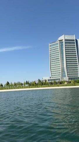 The video features a serene boat ride on a large body of water, likely a lake or pond, under a clear, bright blue sky with a faint, wispy cloud. The camera, mounted on a moving boat, offers a panoramic view of the shore.

Initially, a tall, modern skyscraper, possibly a hotel or corporate building, is visible on the right, with "MINYOUR" clearly displayed near its top. The building has a light-colored facade with numerous horizontal windows.

As the boat progresses and the camera pans left, the shoreline comes into full view, neatly landscaped with rows of green trees and bushes. Beyond these trees, streetlights are visible. Further along the shore, a large, sprawling building with a more traditional architectural style appears. This building features a series of prominent arches along its length, suggesting a public facility or another grand structure.

The water in the foreground is a greenish-blue, showing gentle ripples from the boat's movement. In the final moments of the video, part of a white boat railing and a person's light-colored, wavy hair are visible in the bottom left corner, confirming the vantage point from a moving vessel.

The scene captures a sunny day in what is identified as Kuchtumgali, Uzbekistan, showcasing a blend of modern and traditional architecture along a landscaped waterfront.