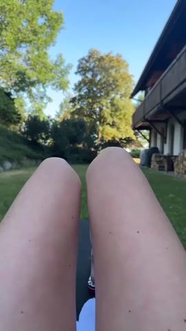 The video provides a first-person perspective of someone lying outdoors, with their legs filling the lower half of the frame. The legs are fair-skinned and appear to be resting on a dark surface, possibly a lounger or mat. Behind the legs, a grassy lawn is visible, with a large tree and other greenery in the background. To the right, a portion of a building with a wooden balcony and stacked firewood is also in view. The sky is clear and blue. The camera appears to be stationary, with subtle movements of the legs causing slight shifts in the image.