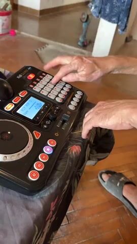 The video shows a close-up view of a person's hands operating a black DJ controller. The controller has a display screen, several buttons, and a large circular jog wheel. The person's hands are actively pressing buttons and manipulating controls on the device. In the background, a wooden floor is visible, along with a table covered with a patterned cloth. Other objects in the background are out of focus, including a power tool and a blue garment hanging on a wall. The lighting suggests an indoor environment.
