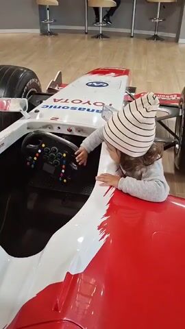 The video shows a young child wearing a striped beanie sitting in a red and white Formula 1 racing car. The child is reaching out and touching the steering wheel, which has various buttons and a small screen. The car is positioned on a wooden floor in what appears to be an indoor setting, possibly a showroom or a play area, with a counter and stools visible in the background.