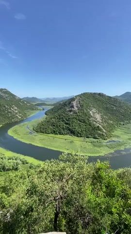A sweeping vista showcases a river meandering through a lush, green landscape under a clear blue sky. The river forms a pronounced U-bend around a verdant, tree-covered hill in the foreground, with its banks lined by bright green vegetation, possibly reeds or algae. In the background, rolling hills extend into the distance, also covered in greenery. The sky above is a brilliant, cloudless blue. The camera slowly pans from left to right, revealing more of the winding river and the expansive natural scenery.
