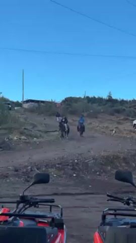 Two horses approach, each ridden by a person. The person on the left is wearing black, while the person on the right is wearing blue. They are coming down a dirt road on an incline. The road is rocky. In the foreground are the front parts of two red ATVs. Overhead are power lines, and in the distance, a building is visible on top of the hill. The sky is clear and blue.