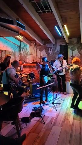 The video captures a live band performing indoors. The group consists of a female vocalist playing guitar at the front, two guitarists, a drummer, and an additional band member barely visible behind the drum kit.  The room is dimly lit with bright pink, blue, and yellow neon lights that accentuate the tropical-themed wall mural featuring painted foliage. The audience includes a woman in the foreground, dancing in front of the band. The setting appears to be a bar or small music venue.