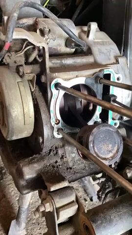 The video shows a close-up view of a partially disassembled motorcycle engine. The engine block is visible, with the cylinder head removed, revealing the piston and crankshaft. Various engine components, wires, and parts are scattered around the engine. The environment appears to be a workshop or garage, with tools and other items in the background. The lighting is somewhat dim, suggesting an indoor setting. There are no people visible in the video. The overall impression is that of a mechanical repair or maintenance operation.