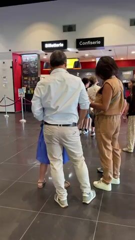 The video opens in a cinema lobby. A man wearing a light blue shirt and beige pants stands with his back to the camera, facing a woman in a tan jumpsuit. To their left, a young girl in a blue dress is partially visible. In the background, there are signs for "Billetterie" (ticket office) and "Confiserie" (confectionery). The floor is tiled in large, dark gray squares.

The camera then pans to the right, revealing a group of people, including children, standing in a line, possibly waiting to purchase tickets or concessions. Red velvet ropes are used to guide the queue. Further to the right, movie posters are displayed on a wall.

The perspective shifts again, moving through the lobby towards another area of the cinema. Two white, wave-shaped kiosks stand prominently in the foreground. Behind them, a digital display board shows movie schedules, and a large movie poster is visible.

The camera continues to move, showing more of the cinema's interior. A large red wall is visible on the left, with a black ventilation grate. To the right of the grate, cardboard cutouts of cartoon characters, possibly from a children's movie, are displayed. A movie poster stands to the right of the characters.

The video ends by panning further right, showing more of the red wall and a colorful display of movie posters in the distance. The overall impression is a bustling cinema lobby with people engaged in various activities related to moviegoing.