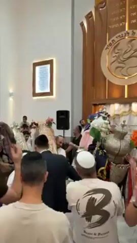 The video shows a lively gathering of people celebrating in what seems to be a synagogue. The room is brightly lit with a high ceiling and adorned with wooden carvings and a framed text on the wall. A group of men, some wearing traditional Jewish head coverings, are carrying what appears to be a Torah scroll covered in a decorative cloth. People are clapping, moving along with the music, and generally participating in a celebratory dance. A speaker stands in the background, presumably amplifying the music.