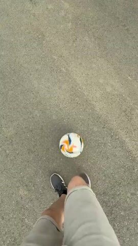 The video shows a person in gray shorts and dark sneakers standing on a gray paved surface with a soccer ball. The person kicks the ball with their right foot, and the ball moves slightly. The camera is positioned above and behind the person, looking down at the scene. The environment appears to be outdoors, possibly a street or a park. The lighting is bright, suggesting daytime. The action is simple and repetitive, with the person kicking the ball back and forth.