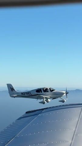 This video shows two airplanes flying in formation against a clear blue sky. The camera appears to be mounted on the wing of one airplane, looking towards another airplane flying alongside. The second airplane is white with the registration number N219EL visible on its fuselage, and the letters "LA" are marked on its tail. The wing of the camera airplane is in the foreground, showing its metallic surface with numerous rivets. The background reveals a hazy landscape of mountains and water, likely an ocean or large lake, under a bright sun. The sun is positioned to the left, casting light on the scene and creating shadows. The overall environment suggests a scenic flight in daylight.