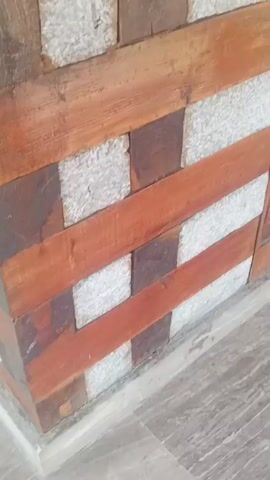 The video shows a close-up view of a wall constructed from wooden planks and rough stone blocks. The wall has a decorative wooden trim on the right side featuring intricate carvings, including what appears to be a figure at the bottom. A metal gate, possibly gold-colored and collapsible, is visible behind the trim. Two pieces of cloth, one yellow and one white, are draped on the gate. The camera pans slightly upwards and to the right, revealing more of the wooden and stone wall construction and a glimpse of an outdoor area with greenery beyond a railing. The lighting suggests daytime.