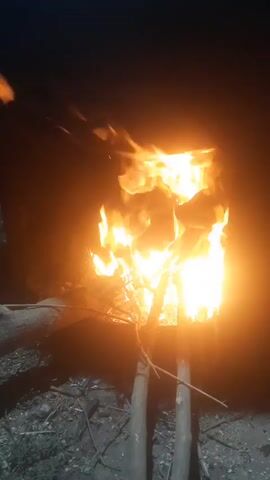 A bright orange fire burns intensely in a dark, outdoor environment. The flames flicker and dance upwards, illuminating the immediate surroundings. The fire is contained within what appears to be a metal structure, possibly a barrel or bin, which is propped up by two wooden sticks or branches. The ground around the fire is covered in dirt, small stones, and twigs. In the foreground, there are also some larger branches or logs lying on the ground. The overall scene suggests a campfire or some form of controlled burning taking place at night. There are no people visible in the video.