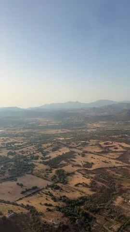 This is a vertical, aerial drone shot of a rural landscape under a clear, pale blue sky. The camera is moving slowly forward and slightly to the right, offering a sweeping view of rolling hills, fields, and scattered trees. The terrain is a mix of dry, golden-brown vegetation and patches of green, suggesting a dry climate or late summer. In the distance, a hazy mountain range forms the horizon. A few small structures, possibly farmhouses or outbuildings, are visible in the foreground and scattered throughout the landscape, appearing as small dots from this altitude. The overall impression is one of vastness and natural beauty.