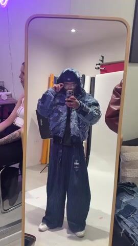 The video shows a person standing in front of a full-length mirror, taking a selfie with a mobile phone. The person is wearing a dark baseball cap, glasses, a blue hooded jacket made from denim patches, a black t-shirt, and loose, blue, pinstriped pants. The person has several tattoos on their hands and neck. 
The room appears to be a studio or workspace. In the background, to the left, a woman is sitting in a chair with a clipboard. Behind the person in the mirror, there's a white backdrop, and what looks like lighting equipment, a red chair, and a leather tote or purse. There is a black couch with pillows to the right of the person, and another denim jacket or garment on the seat of the couch. The room is brightly lit with purple lighting. The camera angle is from the perspective of the mirror's reflection, capturing the entire scene.