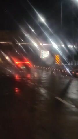 The video captures a nighttime scene on a wet road, likely after rain. A car with red taillights is visible driving away from the camera. The road surface reflects the bright lights of street lamps or overhead lights, creating a shimmering effect. Signs are visible along the side of the road, including a circular sign with the number "60" inside, indicating a speed limit. The overall atmosphere is dark and somewhat blurry due to the conditions and possibly camera movement.