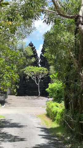 The camera pans up from a street to reveal a Balinese temple. The temple is built of dark stone, with three ornate spires.  A small tree grows in front of the temple. A woman is sitting on a motor scooter near the edge of the frame.  The sky is blue with white clouds. Trees and vegetation surround the temple.