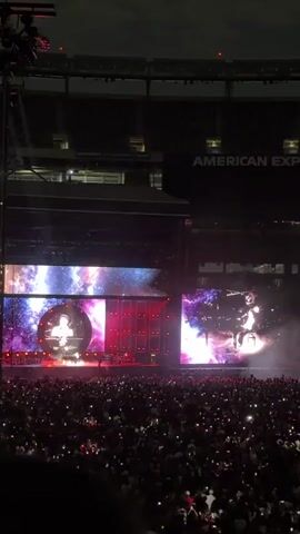 A concert stage is visible with large screens displaying a performer singing at a microphone. The performer is a male with dark, curly hair, wearing a white shirt and a colorful vest. The background on the screens is a nebula or galaxy graphic. Red lights and stage equipment are visible on the left side of the stage. A large audience is in the foreground, with many people holding up their phones with lights on. The stadium seating is visible in the background above the screens, with "AMERICAN EX" visible on a banner. The lighting is dark, typical of a nighttime concert.