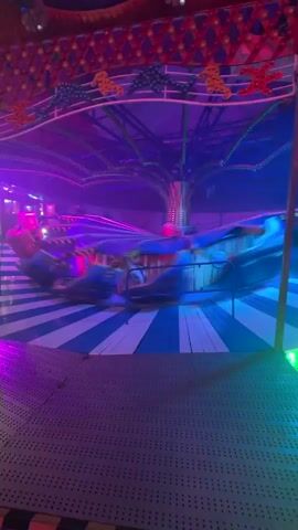 The video shows a carnival ride spinning at night. The ride has a central spinning mechanism with arms extending outwards, each holding a seat or gondola. The ride is brightly lit with red, green, blue, and yellow lights. The floor of the ride is a striped pattern of blue and white. The background is dark and suggests an outdoor setting at night, possibly a fairground. The ride is in motion, creating a blur of color and light. No people are clearly visible, only the movement of the ride itself. The camera is positioned at a low angle, looking up at the spinning ride.
