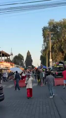 The video captures a lively street scene with a gathering of people and vendors under a clear sky. In the foreground, a man in a white and red outfit walks across the street, gesturing towards the center of the scene. Further ahead, a horse and rider are visible amid a crowd of pedestrians and market stalls sheltered by umbrellas. A silver car is parked on the side of the street, while trees and buildings flank the road, with power lines stretched overhead. The setting appears to be a traditional market or festival.