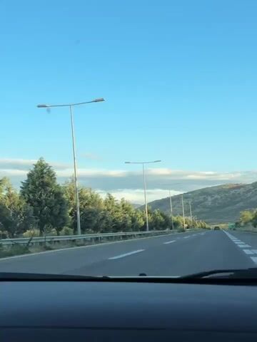 The video shows a view from inside a vehicle traveling on a highway. The sky is a clear blue with some scattered clouds. On the left side of the road, there is a row of evergreen trees and a metal guardrail. Several tall lampposts are visible along the roadside. In the distance, there are rolling hills covered in vegetation. A dark-colored car is seen ahead on the highway. The road has white lane markings. The camera appears to be slightly tilted upwards.