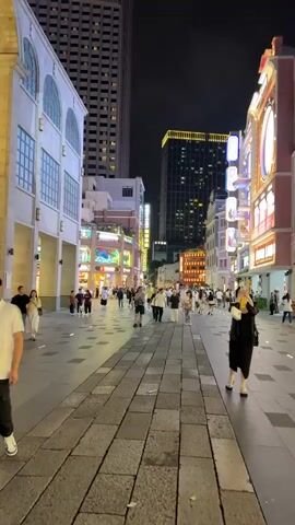 Here's a description of the video:

The video shows a nighttime street scene in what appears to be a bustling commercial district. The street is paved with rectangular stones and is lined with buildings of various architectural styles. Many of the buildings are illuminated with bright neon and LED lights, creating a vibrant and lively atmosphere. There are several tall skyscrapers visible in the background, indicating an urban setting.

Numerous people are walking along the street, moving in both directions. They appear to be a mix of locals and tourists, dressed in casual attire. Some are walking alone, while others are in groups. A woman in a black outfit walks in the foreground, holding a beverage. 

The overall impression is that of a lively and prosperous urban area, with a mix of commercial activity and pedestrian traffic.