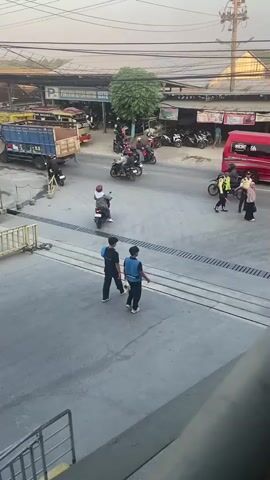 The video shows a busy street intersection with multiple motorcycles and a few cars. Several people are walking or standing, including individuals in what appear to be security or traffic control uniforms. The environment is urban, with buildings and storefronts lining the street. The lighting suggests it is daytime, possibly late afternoon. There are no discernible acts of violence, distress, or emergency. The overall impression is one of normal street activity in a commercial area.
