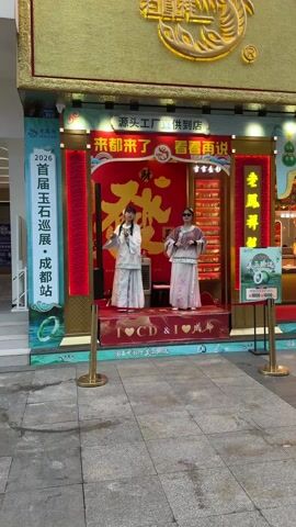 Two young women are dancing on a red platform set up outside of a jewelry store. They are wearing traditional Chinese clothing and dancing to an upbeat song. The jewelry store has large signs written in Chinese. A small air conditioner sits behind one of the women. The platform reads "I [heart] CD & I [heart] Chengdu" in white letters. A red rope is set up to keep people away from the dancers.