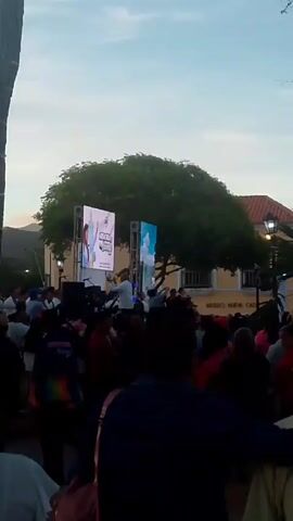 Here is a description of the video:

A large crowd gathers for an outdoor event in front of a yellow building labeled "Museo Nueva Cadiz". A stage is set up with two large screens displaying visuals. A man in white stands on the stage, seemingly addressing the crowd. The sky is a clear blue, and a large tree provides a natural backdrop to the event. The atmosphere appears lively, with the crowd facing the stage. The event is taking place in a public square or park.