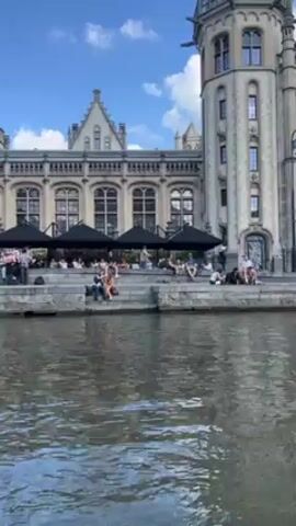 The video depicts a waterfront scene in Ghent, Belgium, likely from a boat traveling on the canal. The camera shows the façade of a grand, old building made of light-colored stone with elaborate architectural details like arched windows and a prominent tower.  A stepped embankment lines the water's edge, where people are relaxing. Some are seated, and a few stand. Several black umbrellas offer shade. The water of the canal reflects the daylight and buildings. The sky is blue with few clouds. The overall atmosphere suggests a leisurely day with people enjoying the beautiful surroundings.