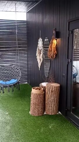 The video begins with a view of a decorated porch.  The walls are dark, and artificial grass covers the floor. A hanging chair with a blue cushion, tree stump stools, and wall decorations of a dreamcatcher and dried plant matter are visible. The camera then pans to an outdoor scene revealing a snow-covered yard with a wooden fence bordering a birch tree forest. The sky is overcast.