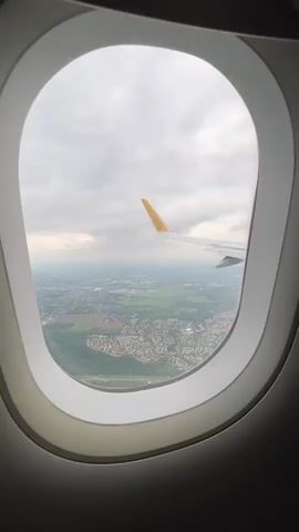 The video captures an aerial view from an airplane window, showcasing the landscape below during flight. A prominent white oval window frame borders the scene. Through the window, the white wing of the airplane is visible, extending from the upper right, with its yellow winglet clearly displaying the text "Vueling."

The ground below, identified as likely over Pontault-Combault, France, presents a mosaic of human development and natural features. Extensive residential areas, characterized by densely packed houses with grey or reddish roofs, are interspersed with large green fields and patches of woodland. A network of roads, including what appears to be a larger highway or main road, crisscrosses the terrain. In the distance, the landscape becomes hazier, suggesting further urban or industrial development.

The sky is heavily overcast with thick, grey clouds, indicating a cloudy daytime environment. The airplane appears to be in a steady flight path, possibly during ascent or descent, as the ground features are quite distinct. A subtle banking movement occurs, causing a slow, gentle shift in the visible landscape. The continuous hum of airplane engines is audible throughout the short clip.