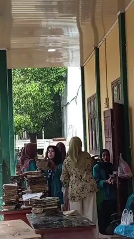 The video shows a group of young women standing on a covered porch. All the women are wearing hijabs and are dressed in clothing that includes a light green color.  They are standing around tables covered in what appears to be stacks of books or papers.  Some of the women are picking up items, possibly distributing materials or taking them away.  The porch has green support beams and a white ceiling.  Behind the women, there is a view of a lush green tree and a building with a crack in the wall.  The overall scene suggests an educational or administrative environment.