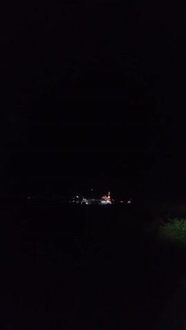 A very dark nighttime video shows a distant illuminated structure, possibly a building or group of buildings, on the horizon. There are some lights visible, with one larger structure appearing to be the main focal point, possibly a church or temple with a tall spire. The rest of the scene is obscured by darkness.