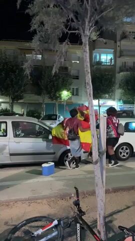 A group of people are gathered around a silver car parked on a street at night. Some of the individuals are holding a Spanish flag. One person appears to be interacting with the car's hood. A blue cooler is on the ground next to the car. A bicycle is lying on its side in the foreground. Buildings with lights on are visible in the background.