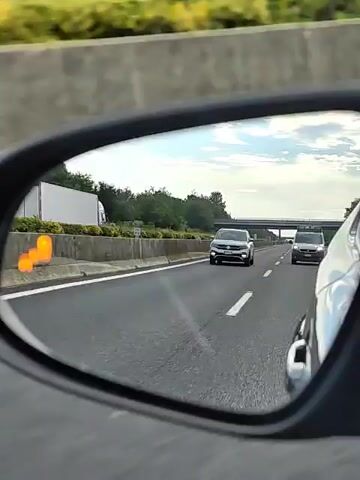 The video shows a view from a car's side mirror while driving on a highway. The mirror reflects the road ahead, with several cars visible. In the foreground of the reflection, a silver Volkswagen SUV is driving in the right lane. Behind it, in the same lane, is a dark-colored SUV. Further ahead, a white semi-trailer truck is visible in the left lane, alongside a concrete barrier. Beyond the truck, more trees and vegetation can be seen. The sky is partly cloudy, suggesting it is daytime.

In the lower left corner of the mirror's reflection, an orange blind-spot warning indicator flashes on the side of the silver Volkswagen SUV. This indicates that a vehicle is present in the car's blind spot. The road appears to be a multi-lane highway. The concrete barrier on the left side of the road is typical of Italian autostradas. There is no visible text in the media file. The overall scene conveys a sense of everyday travel on a highway.