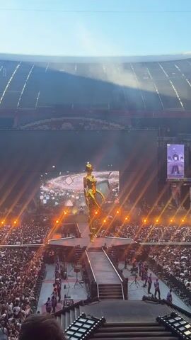 This video captures a live concert taking place in a large stadium, likely at night given the artificial lighting. The central focus is a grand stage with a massive LED screen displaying a cosmic-themed visual. Dominating the stage is a towering, golden statue of a female figure, poised dramatically. Surrounding the stage, thousands of concert-goers are packed together, many with their hands raised, indicating an energetic and engaged audience.

Bright orange lights are strategically placed across the stage, illuminating the statue and the surrounding area, creating a vibrant and theatrical atmosphere. The stadium seating is visible in the background, filled with more spectators. The overall scene suggests a large-scale, popular musical performance. The clear sky and bright sunlight indicate it is daytime, and the stadium looks like it's an open-air venue. No visible text is present in the video.