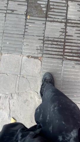 The video, shot from a first-person perspective, shows a person walking on a gray tiled surface. The person is wearing dark-colored clothes, including a long skirt or wide-legged pants with what appears to be a spattered pattern, and dark sneakers. A white decorative accessory is visible on one of the shoes. Occasional dry leaves are scattered on the tiles. At one point in the video, the person makes a "rock on" hand gesture.
