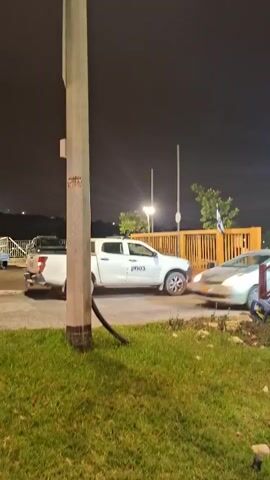 A nighttime scene unfolds with a white pickup truck parked on the left side of the frame. A silver sedan is in motion, passing by the truck. In the background, a wooden fence with a gate stands, and a flag is visible. The ground is covered in grass, and a utility pole is prominent in the foreground. The lighting suggests artificial illumination from streetlights.