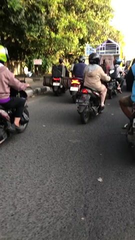 The media file shows a scene of motorbikes on a paved road. The subjects are several individuals riding motorbikes, most wearing helmets in various colors including green and black. One person in the foreground is wearing a light brown jacket and has a black helmet. The scene depicts a procession of motorbikes, suggesting a form of transportation or possibly a convoy.

The setting appears to be outdoors, with lush green trees and foliage lining the side of the road. The presence of what looks like palm trees in the background suggests a tropical or subtropical environment. The road surface is asphalt, and there are some markings on it. In the distance, there's a glimpse of a building with a blue roof. The time of day appears to be late afternoon, judging by the warm, golden sunlight casting shadows and illuminating the scene. The weather seems pleasant, with the sun shining.

There are no overt emotional expressions visible due to the distance and the subjects' focus on riding. The activity is primarily movement along the road, with the motorbikes in motion. One motorbike in the foreground has a white and blue design. Another motorbike further back has a black box attached to its rear. License plates are visible on some of the motorbikes, with one clearly showing "N 5031 JLZ". Another license plate is partially obscured.

A sign is visible on a pole near the trees, but the text is illegible. The overall impression is a common scene of daily life and transportation in a populated area, likely in a region where motorbikes are a primary mode of travel. The location context provided is Klurak, Indonesia, which aligns with the visual cues of tropical vegetation and the style of motorbikes and attire.