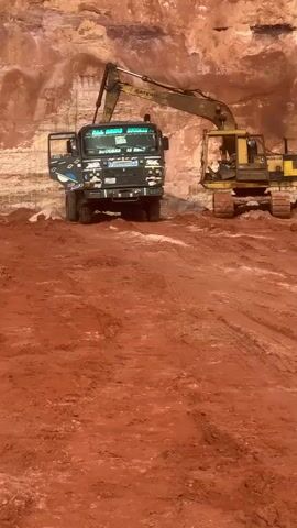 The video shows an excavator loading dirt into a dump truck in a quarry. The excavator is yellow and has a long boom. The dump truck is black and has a blue and white graphic on the side. The quarry walls are made of red dirt. The ground is covered in red dirt. The video is shot from a low angle.