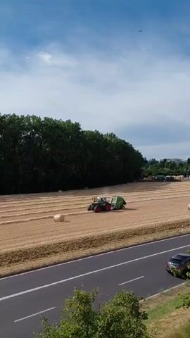 This media file shows a rural scene in Ecublens, Switzerland, on a sunny day with scattered clouds. The primary subject is a green tractor operating in a harvested field, likely baling straw. A large, round hay bale is visible near the tractor, and several other bales are scattered across the field, indicating the baling process is underway. The field is bordered by a dense line of tall, dark green trees.

In the foreground, a highway with two lanes of traffic runs horizontally across the frame. A motorcycle is seen riding on the highway, and further down the road, a white truck is visible. The sky is bright blue with fluffy white clouds, and a bird is soaring high above. The overall atmosphere is peaceful and rural, suggesting a typical agricultural activity during a clear day. The time of day appears to be late morning or early afternoon due to the bright sunlight and clear shadows. No visible text is present in the media file.