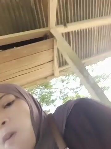 A video shows two people sitting inside a structure with a wooden roof. In the foreground, on the left, a young girl wearing a brown hijab and a white outfit is looking at the camera and smiling. She raises her hand, which is partially obscured. Next to her, on the right, is a man wearing a white t-shirt with a black star logo. He is looking down and appears to be talking or interacting with the girl. Between them, there is a mat on the ground and some food in a clear container. The background shows trees and what appears to be an outdoor environment. The camera movement is somewhat shaky.