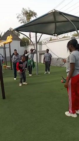 A group of about 10 people are on a green artificial turf playground. One person in red striped pants is holding the end of a long jump rope that goes to a person in a red hoodie who is jumping over it. Most of the other people are standing and watching, but one person off to the side in a blue track suit is jumping as well. The playground has a gazebo-like metal structure, with a yellow decorative piece that looks like a hand. A white wall and trees are visible in the background.