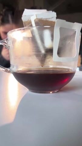 A close-up shot shows a clear glass mug partially filled with dark liquid, likely coffee, on a white surface. Two pour-over coffee filter packets are perched on the rim of the mug. Sunlight illuminates the mug and creates a shadow on the table. The background is blurred, showing a person with their hair up, possibly using a phone. There is a brief sound of a stifled laugh.